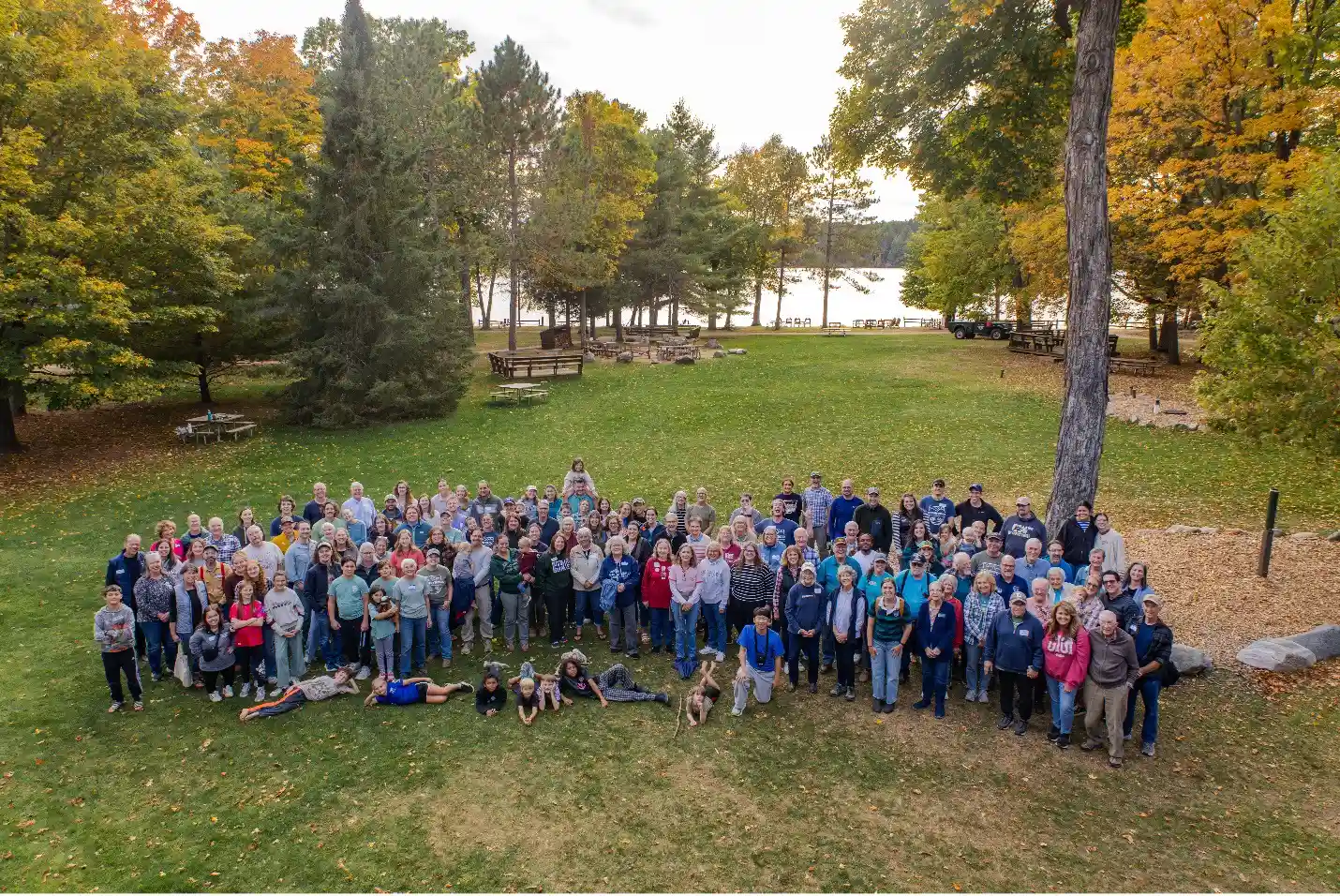 A large group gathers on a grassy lawn beneath autumn trees, with a lake behind them