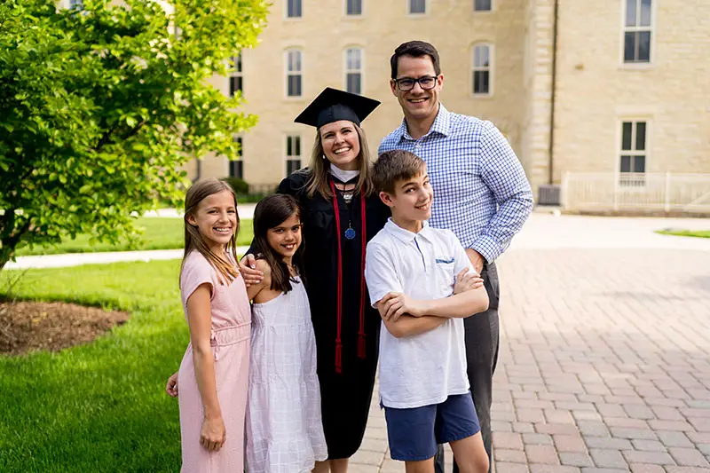 A smiling graduate in cap and gown poses with a man and three children on a sunny campus