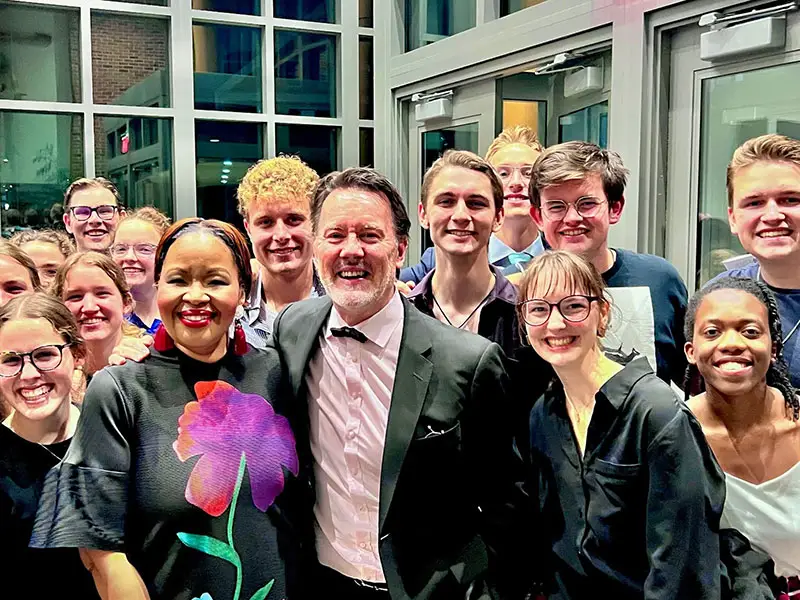 A smiling man in a suit and woman in a flower-print top pose with a group of young people in a building