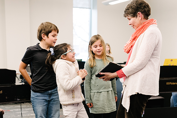 A teacher is holding a tablet, engaging with three children.