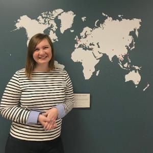 Maddie MacMath standing in front of Intercultural Studies department hallway map and smiling