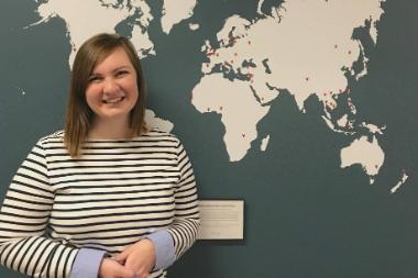 Maddie MacMath standing in front of Intercultural Studies department hallway map and smiling