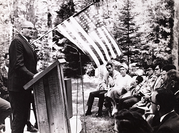 A man speaks at a wooden podium outdoors, with an audience seated under trees with an American flag by the side