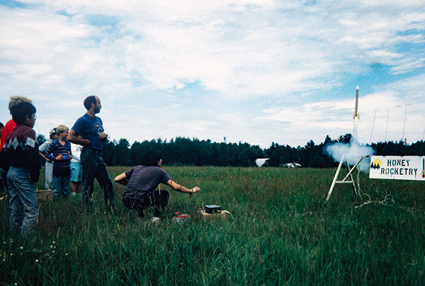 A group of people watch a model rocket launch in a grassy field