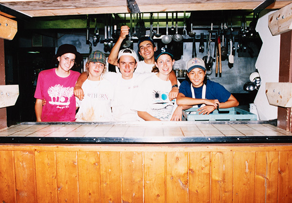 A group of young people stand behind a wooden kitchen counter, smiling