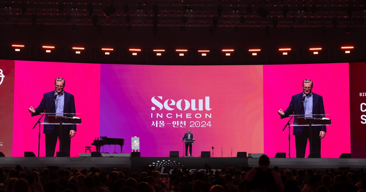 A white man stands on a stage in front of a large pink screen on which he is projected