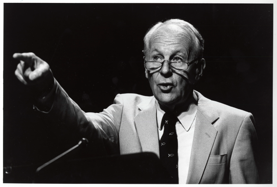 A black and white photo of a white-haired man with glasses speaking from a podium
