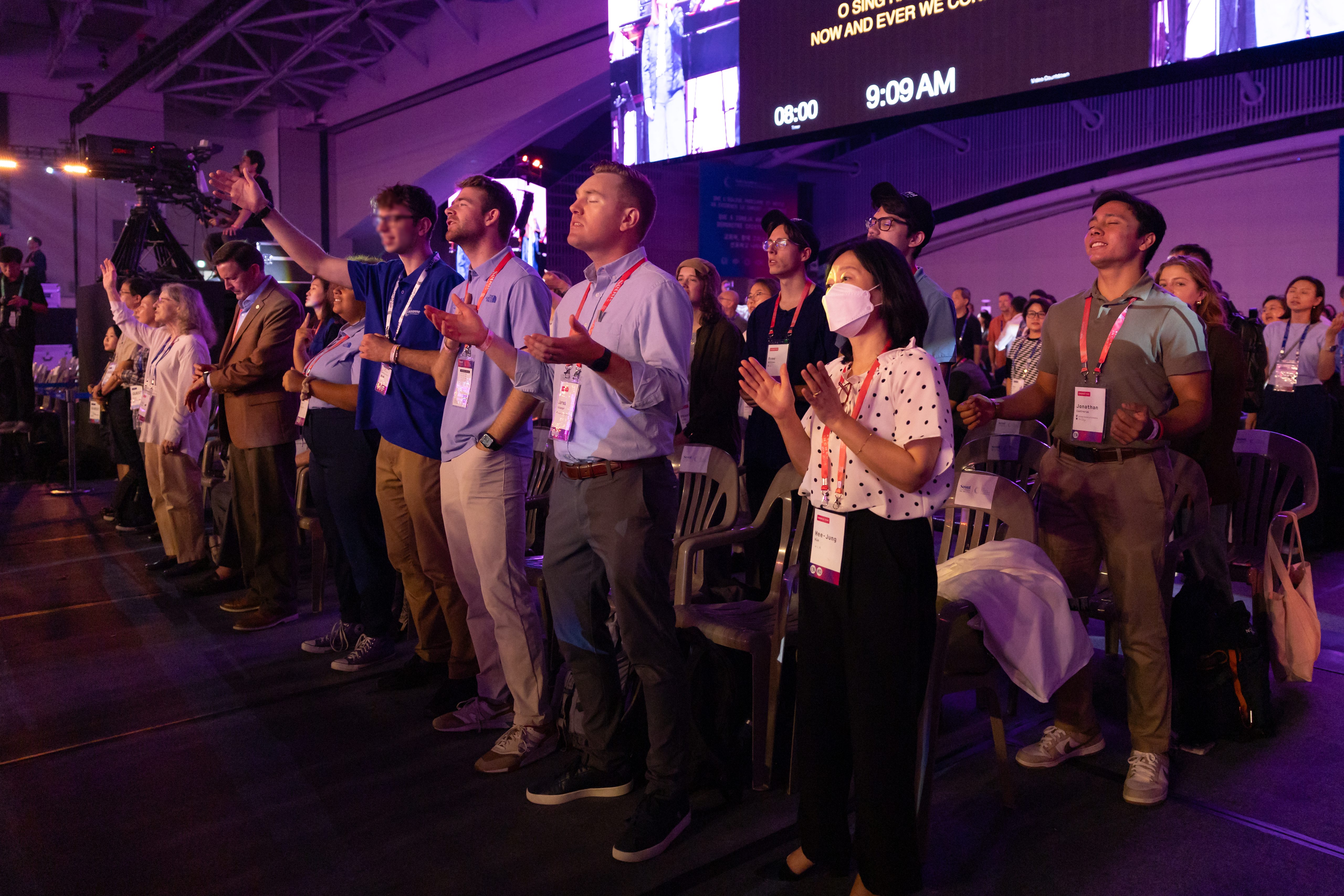 Several young adults stand with two adults in audience and are singing. Many of them are raising their hands in worship.ny of them raise their hands
