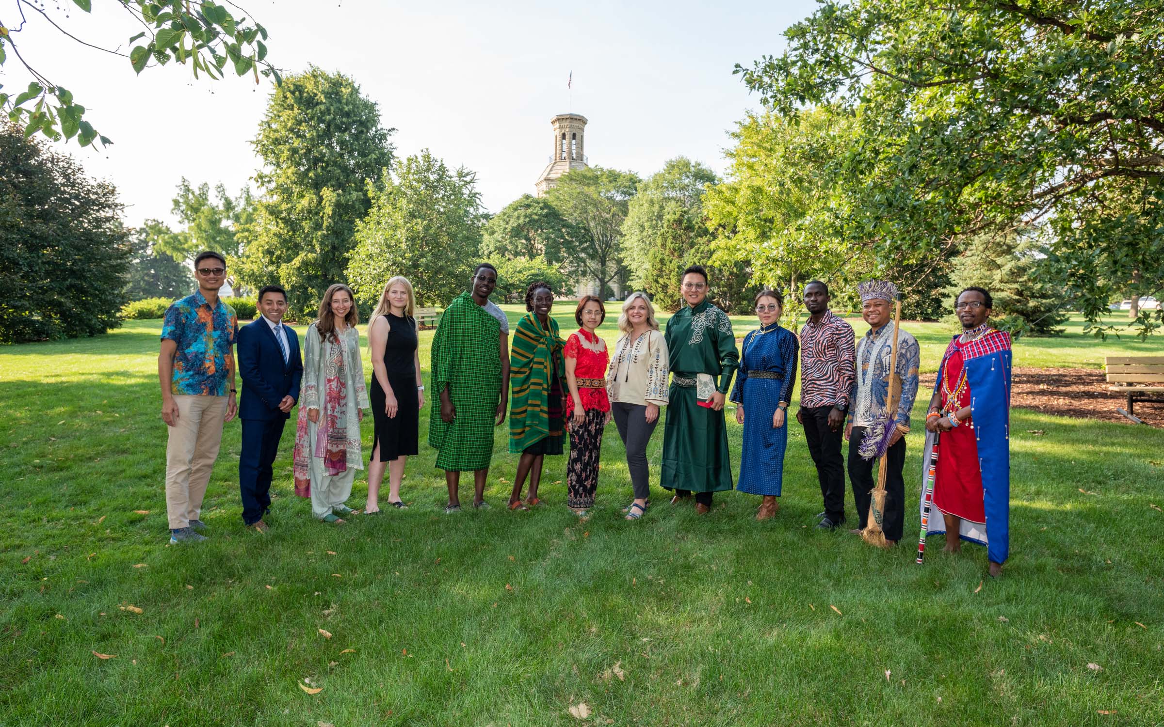 A group of adults from various countries wear traditional clothing from their country and smile for a photo on Wheaton College's campus