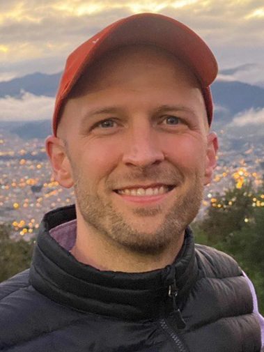 A white man wearing a dark jacket and orange baseball cap smiles