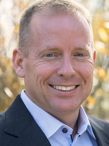 A white man with blue eyes stands outside smiling