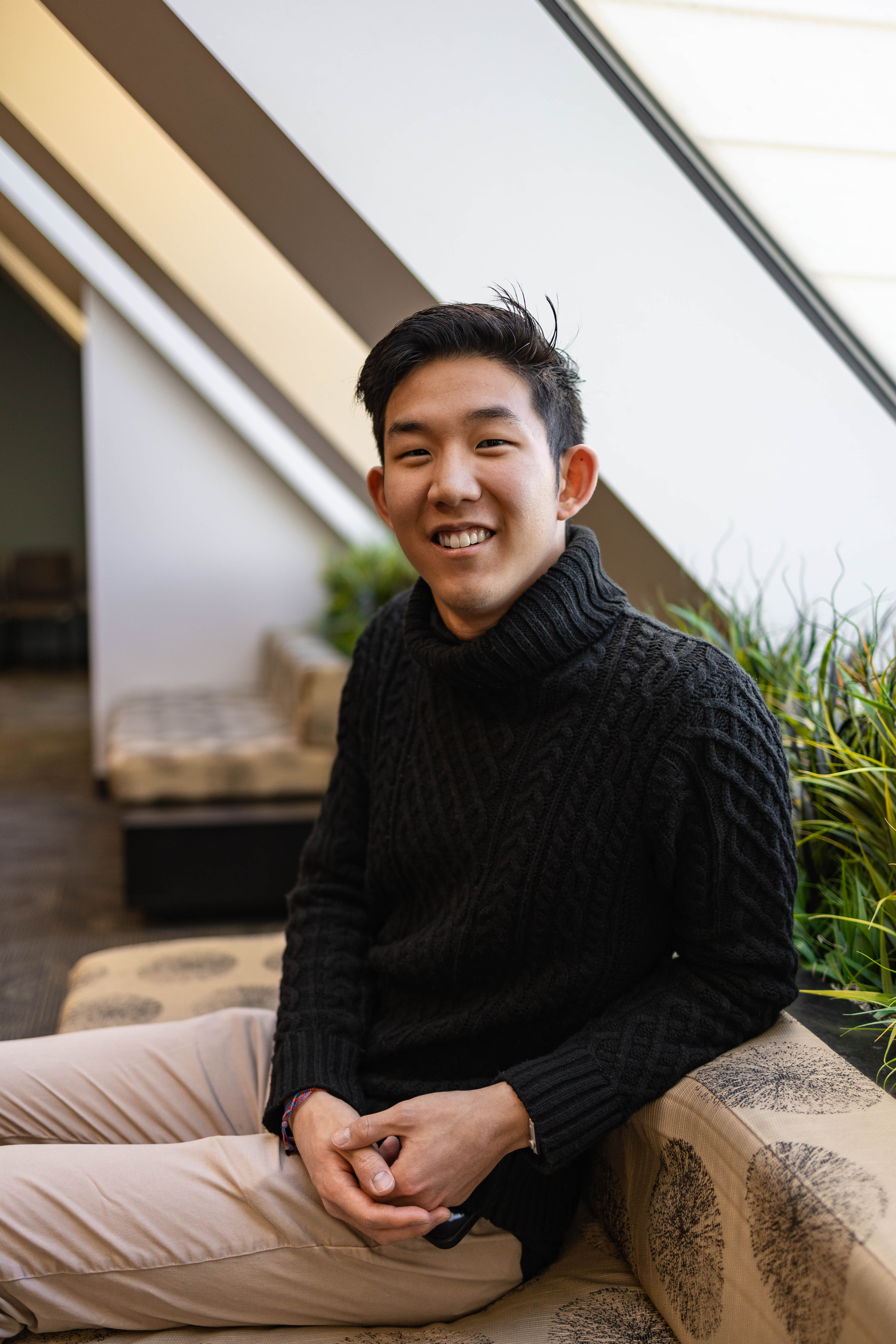 A young East Asian male sitting by a window and wearing a black sweater smiles