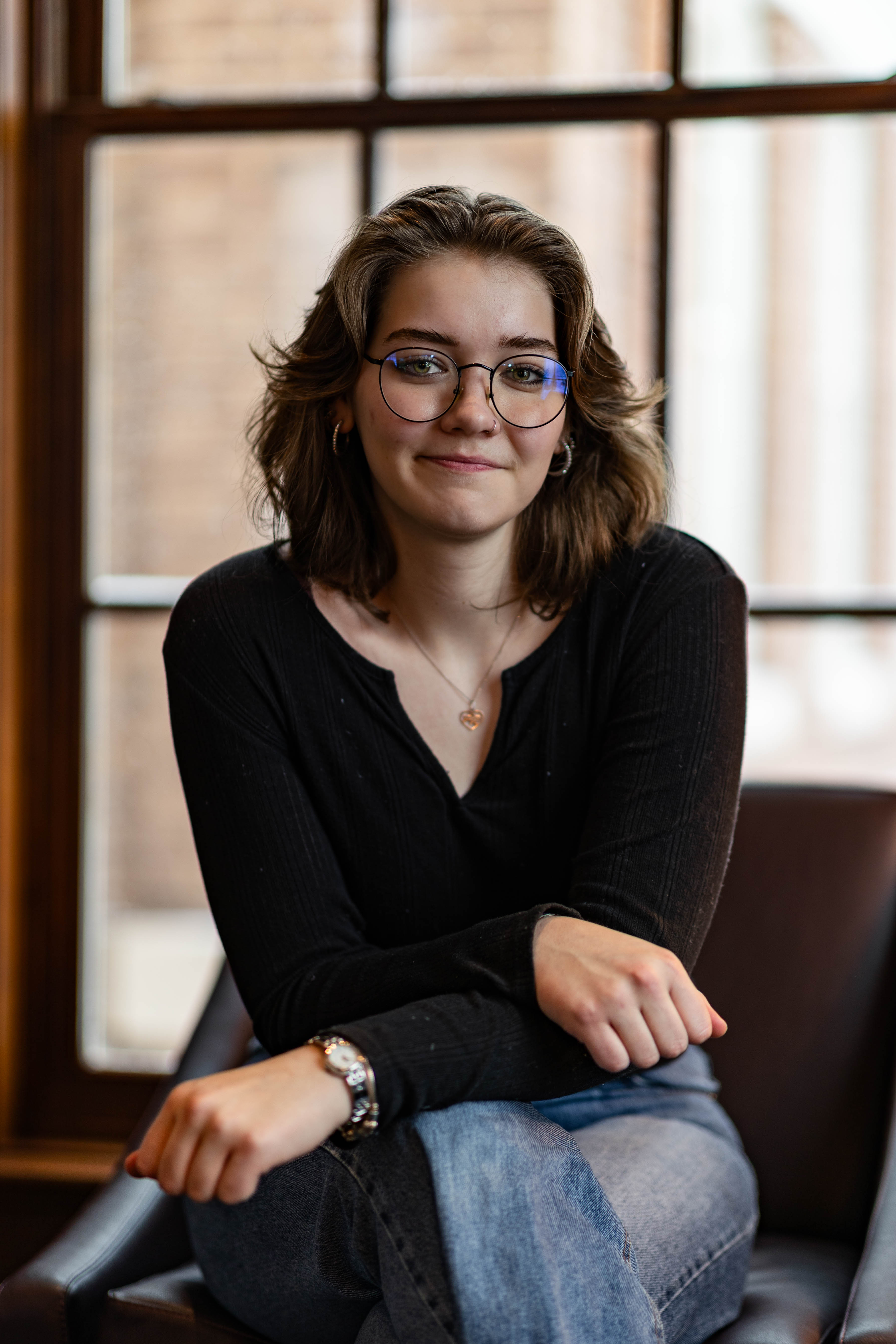 A young white female sitting by a window and wearing a black sweater smiles