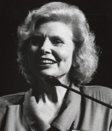 A black and white photo of a white woman smiling and standing at a podium