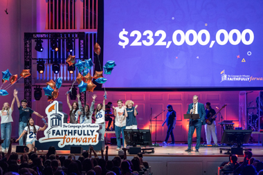 Numerous people stand on a stage holding a sign that reads Faithfully Forward while the screen behind them reads two hundred and thirty two million dollars