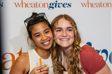 An east asian young woman and a white young woman smile in front of a backdrop that says Wheaton Gives on it