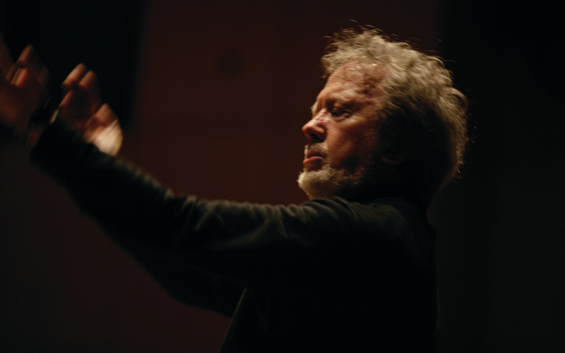 A gray haired white man conducts using his hands