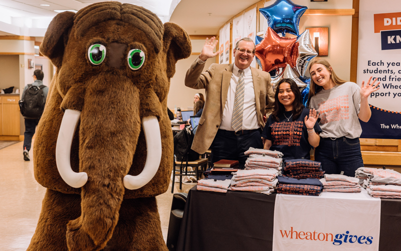 Two female college students and a male college president stand next to a mastodon mascot